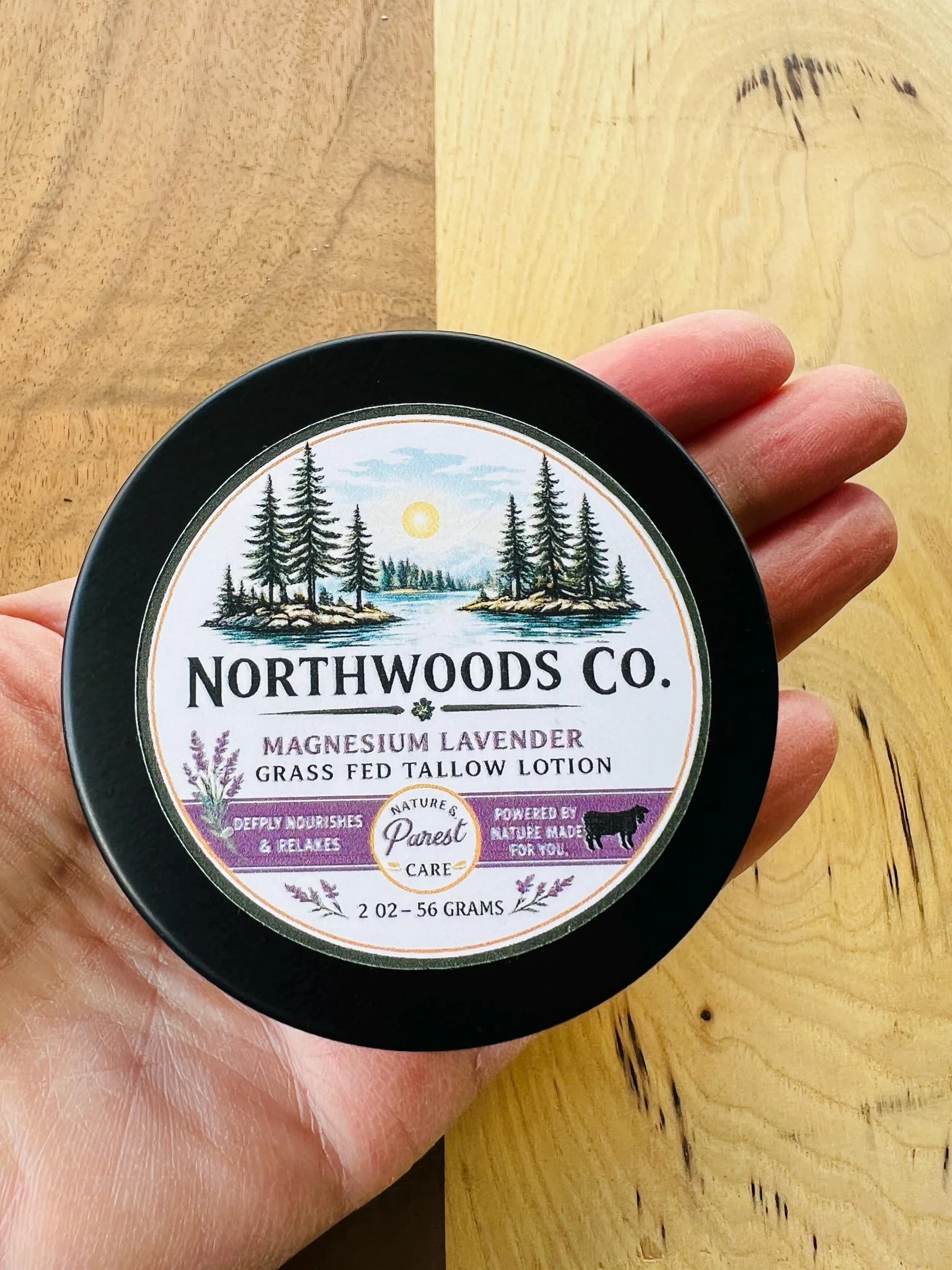May include: A black, round container of Northwoods Co. Magnesium Lavender Grass Fed Tallow Lotion. The label features a scenic illustration of a lake and pine trees. The container holds 2 oz - 56 gra